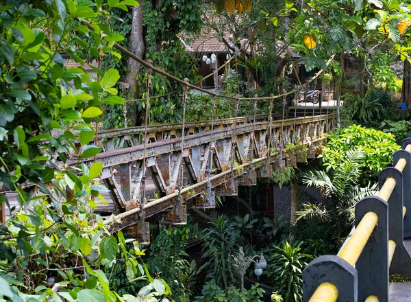 Old iron bridge with wood planks in middle of the rainforest in Bali, Indonesia. Tropical foliage surrounding and motorcycle parked in distance.