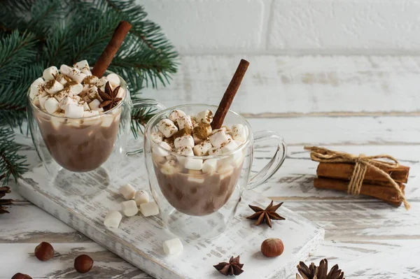 chocolate drink in transparent mugs with marshmallows, cinnamon sticks and anise stars on a white wooden background opposite the brick wall