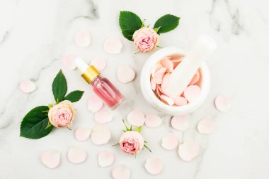 Bir şişe gül yağı, tomurcuklar ve çiçek yaprakları, havan topuzu ve gül yapraklarıyla kompozisyon. Mermer beyaz arka plan. en üstteki görünüm