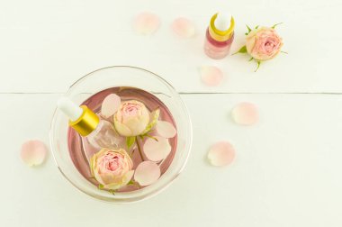 Gül yapraklarıyla dolu bir kasede pembe kozmetik suyu, beyaz arka planda gül özü olan damlatıcı şişeler. en üstteki görünüm