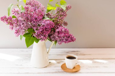 Tahta beyaz bir masada beyaz bir sürahi içinde bahar mor çiçekleri. Bir fincan sabah kahvesi. Ülke eğlencesi kavramı