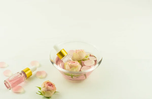 Dwie szklane butelki kosmetyczne z olejkiem różanym, pączkami i płatkami róż w szklanej misce na białym stole. — Zdjęcie stockowe