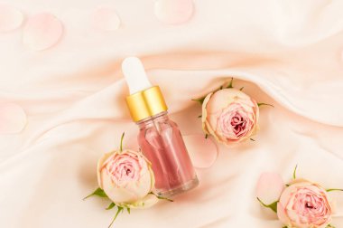 Cilt bakımı için pembe ya da olmazsa olmaz yağlı damlacıklı cam şişe tomurcukları ve çiçekleri olan pembe bir tekstil arka planında.