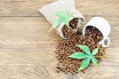 a composition of a coffee cup filled with coffee beans and a bag of coffee beans on a brown wooden background with green morijuana leaves. top view