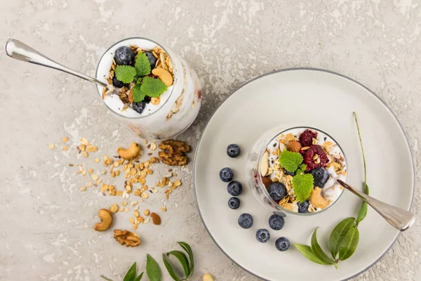 两杯美味的天然酸奶和意大利面 新鲜的浆果 坚果和薄荷叶在灰色的混凝土背景上 顶部视图 — 图库照片