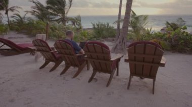 Ortalama 4K 'lık serbest gezgin erkek fotoğrafı. Güzel okyanus kıyısında, gün batımı ve avuçlarında dizüstü bilgisayarıyla çalışıyor.