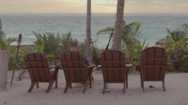 Ortalama 4K 'lık serbest gezgin erkek fotoğrafı. Güzel okyanus kıyısında, gün batımı ve avuçlarında dizüstü bilgisayarıyla çalışıyor.