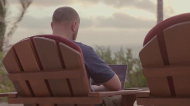 Ortalama 4K 'lık serbest gezgin erkek fotoğrafı. Güzel okyanus kıyısında, gün batımı ve avuçlarında dizüstü bilgisayarıyla çalışıyor.