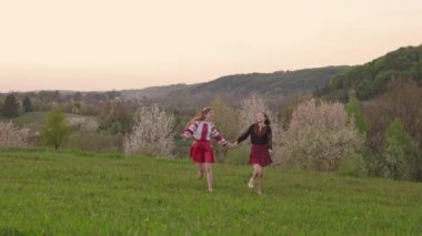 wide 4k shot of two girls in traditional ukrainian vysyvanka holding hands and running in the field during sunset.