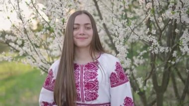 Vyshyvanka 'lı güzel Ukraynalı bir kadının 4k pozu - Ukrayna ulusal kıyafetleri. Çiçek açan ağacın yanındaki kırsalda bir kadın..