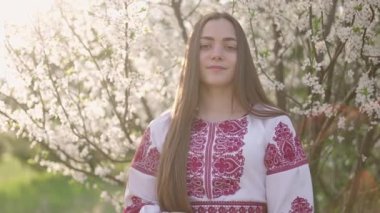 Vyshyvanka 'lı güzel Ukraynalı bir kadının 4k pozu - Ukrayna ulusal kıyafetleri. Çiçek açan ağacın yanındaki kırsalda bir kadın..