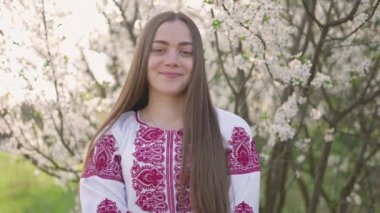 Vyshyvanka 'lı güzel Ukraynalı bir kadının 4k pozu - Ukrayna ulusal kıyafetleri. Çiçek açan ağacın yanındaki kırsalda bir kadın..