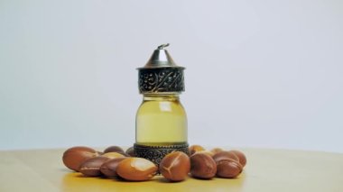 Doğulu cam ve metal bir şişede Argan yağı ve tahta masa ve beyaz arka planda argan fındığı hareketi.