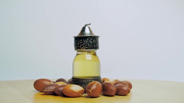 Aceite de argán en una botella de vidrio oriental y metal y el movimiento de las nueces de argán en la mesa de madera y fondo blanco. — Vídeo de stock