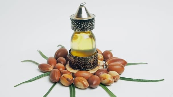 Olio di Argan in bottiglia orientale di vetro e metallo e noci di argan con foglie verdi movimento su sfondo bianco. — Video Stock