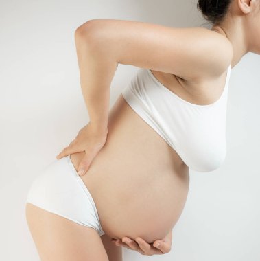 Hamileliğin üçüncü üç ayındaki bir kadın. Beyaz iç çamaşırlı hamile bir kadın karnını tuttu ve belinin alt kısmını beyaz arka planda tuttu. Hamileliğin son ayındaki kız sırtında ağırlık ve gerginlik hissediyor..