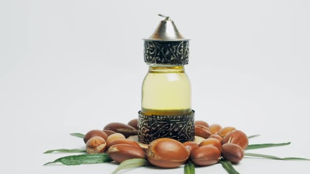 Aceite de argán en una botella oriental de vidrio y metal y nueces de argán con hojas verdes en movimiento sobre fondo blanco. — Vídeo de stock