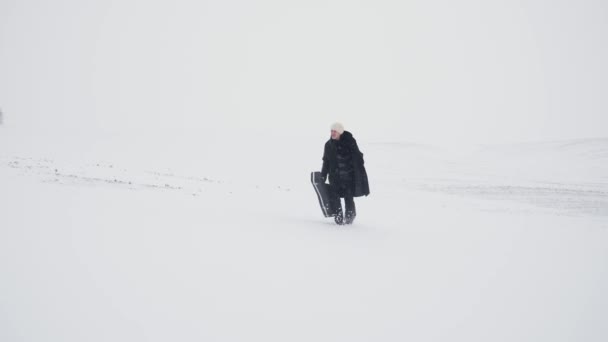 Dorosły mężczyzna w czarnym płaszczu z pudełkiem na gitarę chodzi twardo po zaśnieżonym polu. — Wideo stockowe