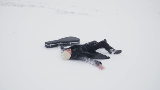 Een volwassen man met een gitaardoos ligt in een sneeuwstorm en probeert op te staan. — Stockvideo