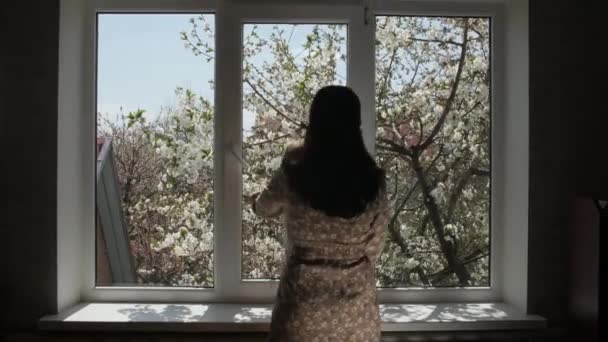 Una mujer con un bebé se acerca a una ventana de metal-plástico. Los cerezos florecen fuera de la ventana. — Vídeo de stock