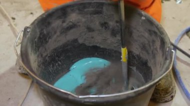 Mavi ve gri fayans yapıştırıcısı siyah kovada elektrikli matkapla karıştırılır. Bu da turuncu tulumlu bir işçinin ayakları tarafından klemplenir..