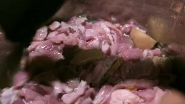 a black-gloved cook stirs fresh pork pieces and melted lard with a spoon in a stainless steel pan. close-up the camera does not move.