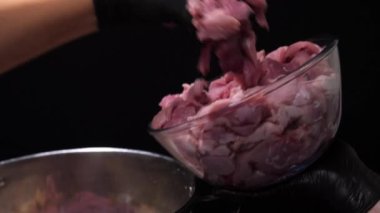A cook in black gloves transfers raw pork from a glass bowl to a stainless steel pan. static general frame from the side.