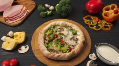 Presentation of pizza on a dark brown table with its ingredients. There are chopped ham, paprika, mushrooms, and cheese. Creamy sauce in a glass.
