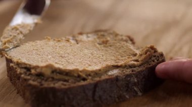 Spread a slice of bread with pate with a stainless steel table knife. The action takes place on the background of a wooden board. A persons fingers support a sandwich.