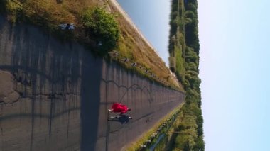 Dikey video. Genç bir çift, bir erkek ve bir kadın, yol boyunca koşuyorlar. Gri takım elbiseli damat, kırmızı elbiseli gelin. Solda büyük bir göl, sağda bir çayır var..