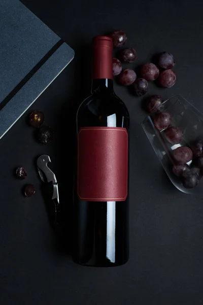 Wine bottle with glass, grapes, bottle opener and notebook, dark background