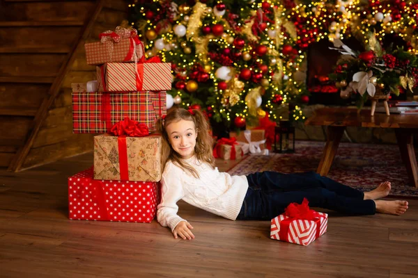 Портрет молодой девушки, сидящей у елки с большой грудой упакованных подарочных коробок, счастливой, получившей много подарков в день бокса. Гора даров падает вместе с ребенком. Шаг 5 Стоковое Изображение
