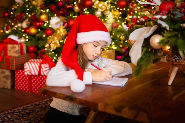 Счастливого Рождества и счастливых праздников. Маленькая девочка в кепке Санта-Клауса пишет письмо Санта-Клауса и мечтает о подарке фоне новогодней елки в помещении. Открытки, Рождественское настроение. Стоковая Картинка