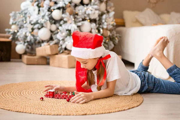 Счастливый ребенок в красной шляпе играет с игрушечным древесным поездом под елкой Лицензионные Стоковые Изображения
