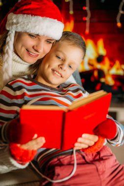 Yeni yıl şapkalı bir anne, oğluna şöminenin arkasında renkli bir kazak giymiş bir kitap okur.
