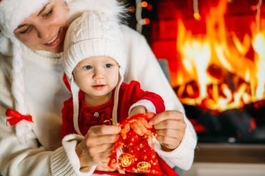 Noel şapkalı bir anne, şöminenin arka planında kırmızı elbiseli, şenlikli bir kızı gösteriyor.