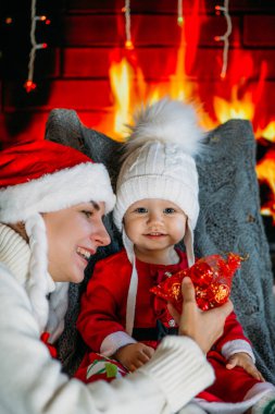 Noel şapkalı bir anne, şöminenin arka planında kırmızı elbiseli, şenlikli bir kızı gösteriyor.