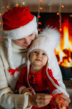 Yeni yıl şapkalı bir anne, kırmızı elbiseli bir kızı şöminenin arka planında tutuyor.
