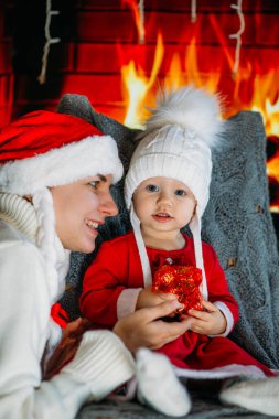 Noel şapkalı bir anne, şöminenin arka planında kırmızı elbiseli, şenlikli bir kızı gösteriyor.