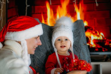 Noel şapkalı bir anne, şöminenin arka planında kırmızı elbiseli, şenlikli bir kızı gösteriyor.