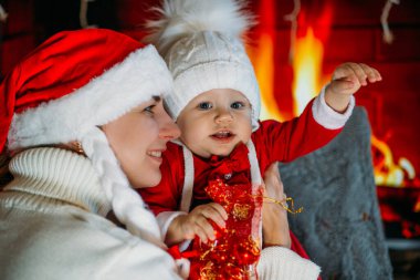 Yeni yıl şapkalı bir anne, kırmızı elbiseli bir kızı şöminenin arka planında tutuyor.