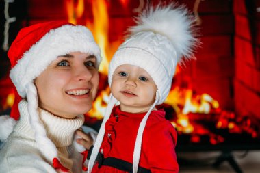 Yeni yıl şapkalı bir anne, kırmızı elbiseli bir kızı şöminenin arka planında tutuyor.