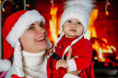 Yeni yıl şapkalı bir anne, kırmızı elbiseli bir kızı şöminenin arka planında tutuyor.