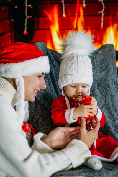Noel şapkalı bir anne, şöminenin arka planında kırmızı elbiseli, şenlikli bir kızı gösteriyor.