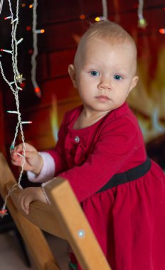 One-year-old girl in red Christmas dress on blurred background of garlands and fireplace
