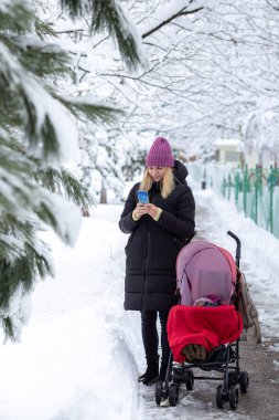 Bebek arabasının yanında genç bir anne. Ve gülümsüyor. Telefondan karlı bir parkın arka planına doğru dönüyor.