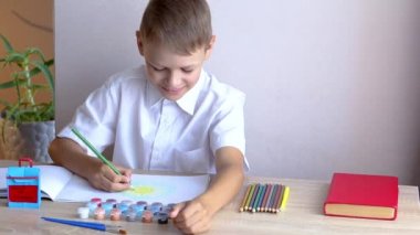 Beyaz tişörtlü bir okul çocuğu okul tabelasının arka planındaki albümde güneşi çiziyor..