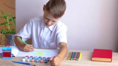 Beyaz tişörtlü bir okul çocuğu okul tabelasının arka planındaki albümde güneşi çiziyor..