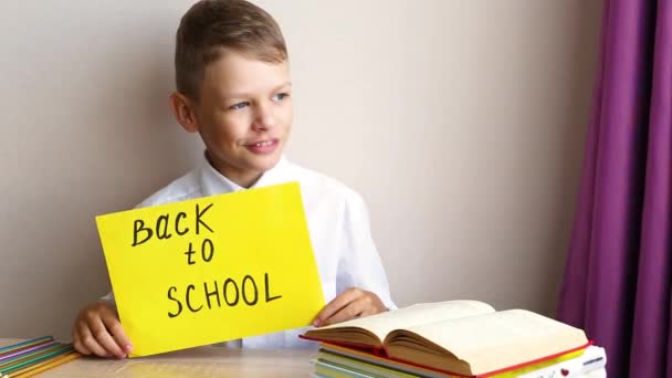 Happy schoolboy dengan kemeja putih duduk di meja berlawanan dengan latar belakang buku teks dengan tanda kuning kembali ke sekolah — Stok Video