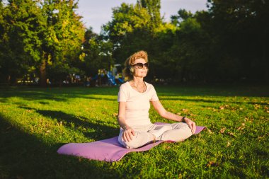 Parkta yoga. Nilüfer çiçekli orta yaşlı kadın yeşil çimlerin üzerinde yoga minderinde oturuyor. Sakinlik ve meditasyon kavramı. Yoga açık havada. Kıdemli kadın doğada meditasyon yapıyor ve yoga nilüfer pozisyonu uyguluyor..